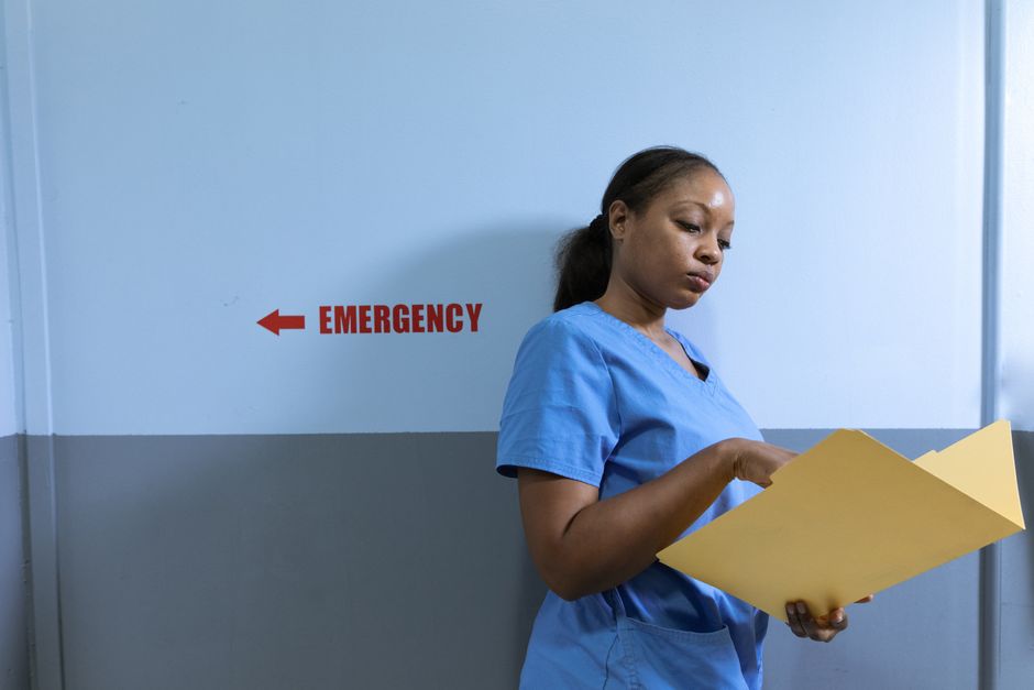 A nurse in blue scrubs reads a folder near an emergency room sign in a hospital.