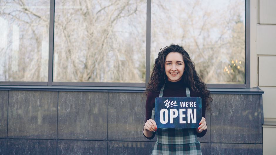 Happy woman holding 'Yes We're Open' sign in front of a store.