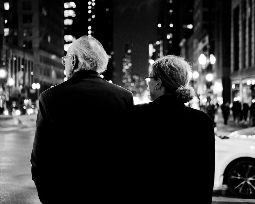 Black and white photo of an elderly couple walking together through a bustling city street at night.
