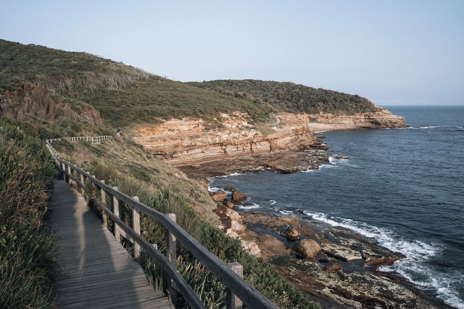 Wooden boardwalk along the scenic coast of Sydney, Australia, with rocky cliffs and the ocean.